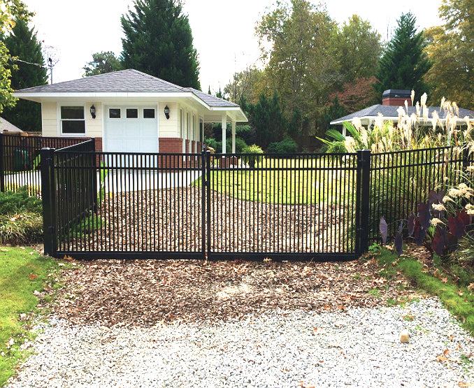Black metal aluminum driveway gate for pets and children security and fence in front of a house with trees and a driveway.