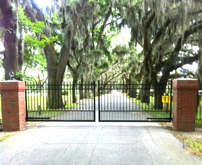 Black metal automated gate with puppy pickets and brick pillars in a park-like setting with trees and Spanish moss.